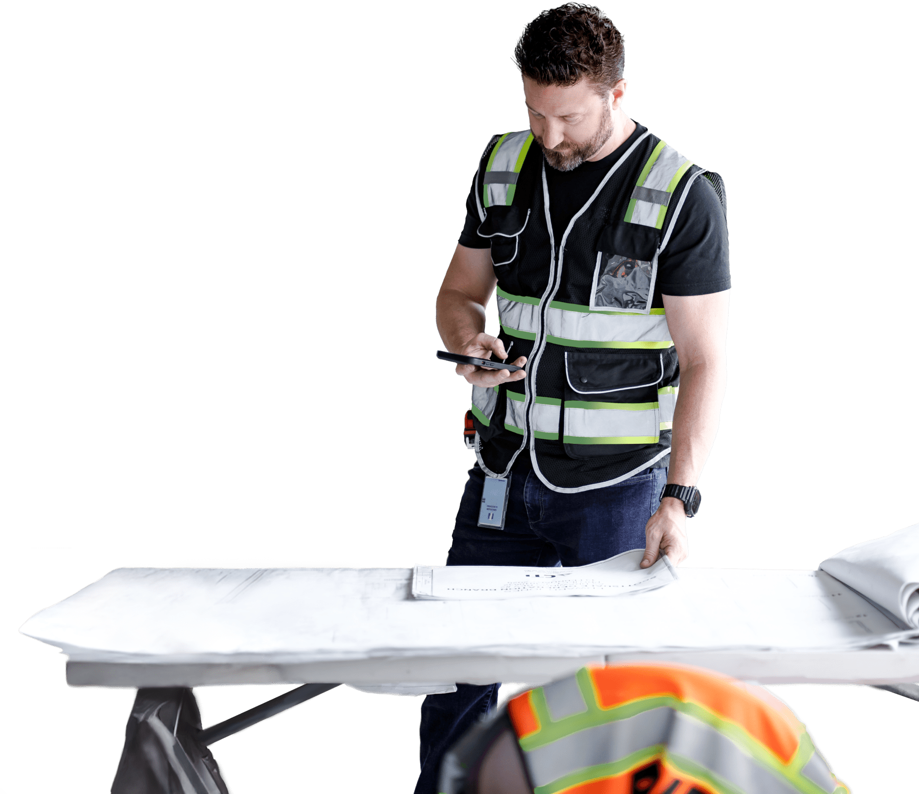 A construction worker in a safety vest looks at a smartphone while standing at a table with blueprints on a commercial construction Arizona site; another worker in a helmet is partially visible in the foreground.