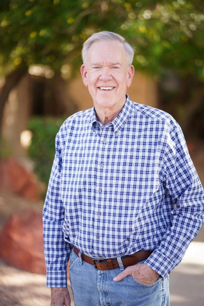 An older man with gray hair wearing a blue and white plaid shirt and jeans stands outdoors, smiling at the camera. Trees and rocks are visible in the background.