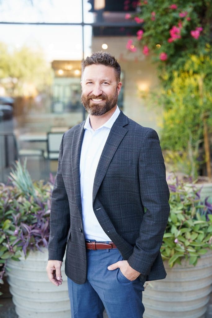 A man with a beard, wearing a blazer and dress shirt, stands outdoors in front of plants and a glass building, smiling at the camera.
