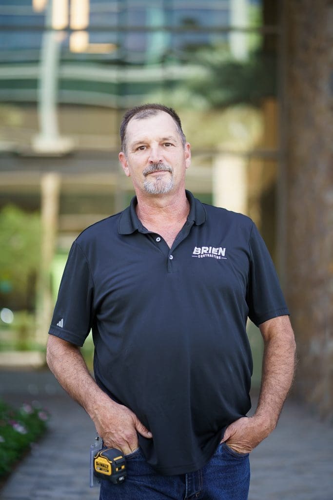 A man in a black "O'Brien" polo shirt stands outdoors with one hand in his pocket and a tape measure clipped to his jeans.