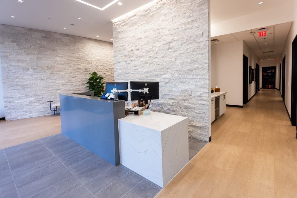 front receptions area for law firm with a large desk holding two monitors with a white rock wall behind and a plant.