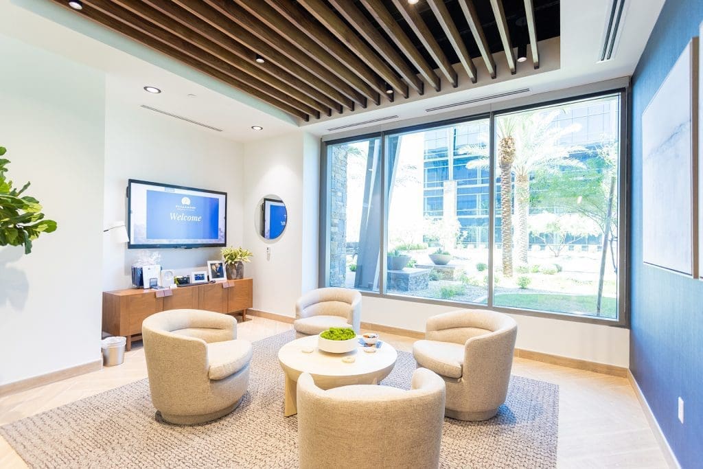 Modern office lounge with four beige chairs around a round table, large window with garden view, wall-mounted TV, and wooden ceiling accents—expertly crafted through top-tier general contracting services in Arizona.
