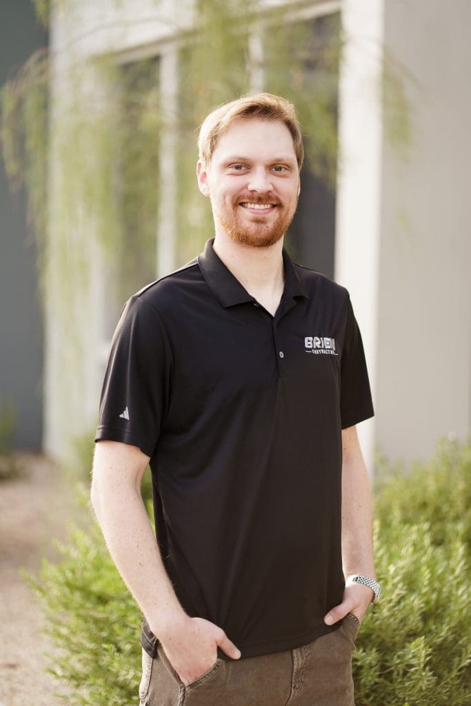 A man with light hair and a beard stands outdoors, smiling, wearing a black polo shirt with "BRI" text on it, with greenery and a building in the background.