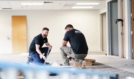Two men in work shirts measure materials and install flooring in a partially finished, empty room with open doors and overhead lighting—typical of commercial construction jobs in Arizona.