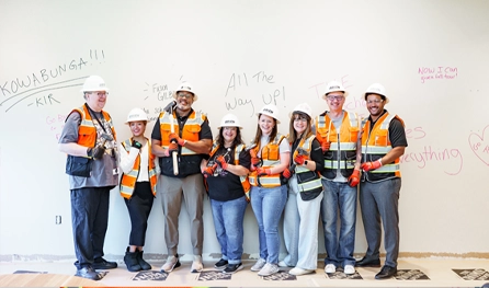 Eight people wearing construction safety gear and vests stand in a row, holding tools, showcasing the teamwork behind commercial construction jobs in Arizona, in front of a wall with messages and drawings written on it.