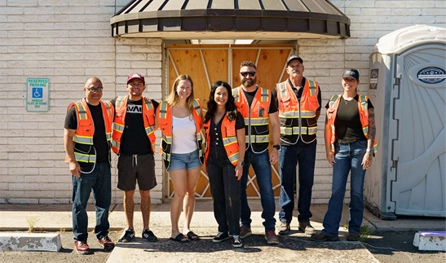 Seven people wearing orange safety vests stand in a row outside a building, with a porta potty visible on the right—a typical scene on commercial construction jobs in Arizona.
