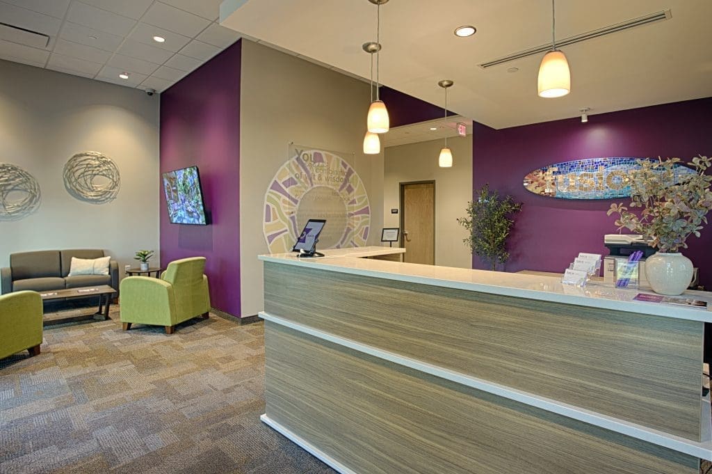 Entry for school with a large wood desk with white countertop, purple accent walls, green chairs, mosaic surfboard Fusion signage and a mosaic fusion mission signage on the walls.