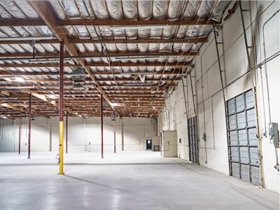 Interior of an empty warehouse with concrete floors, high ceilings with exposed beams, and large roll-up metal doors along one wall.