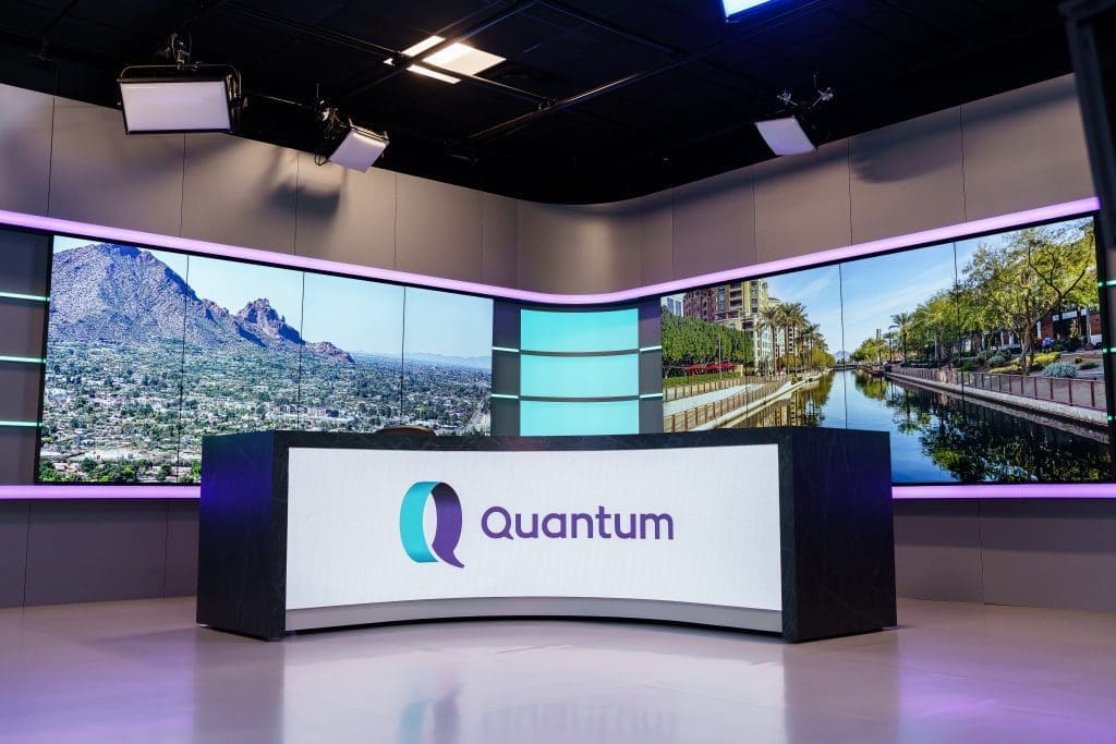 A rounded news desk with a light upfront of branding Quantum. Behind the desk are two large screens for videos or photos and purple light in the background.