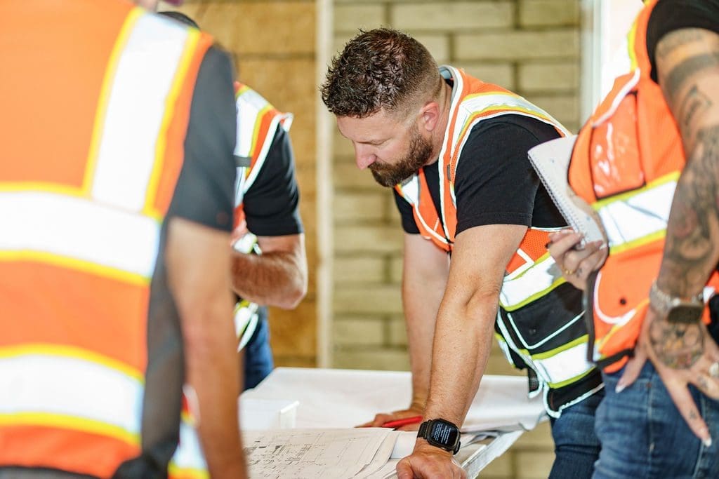 A group of construction workers wearing safety vests review blueprints on a table inside a building under construction.