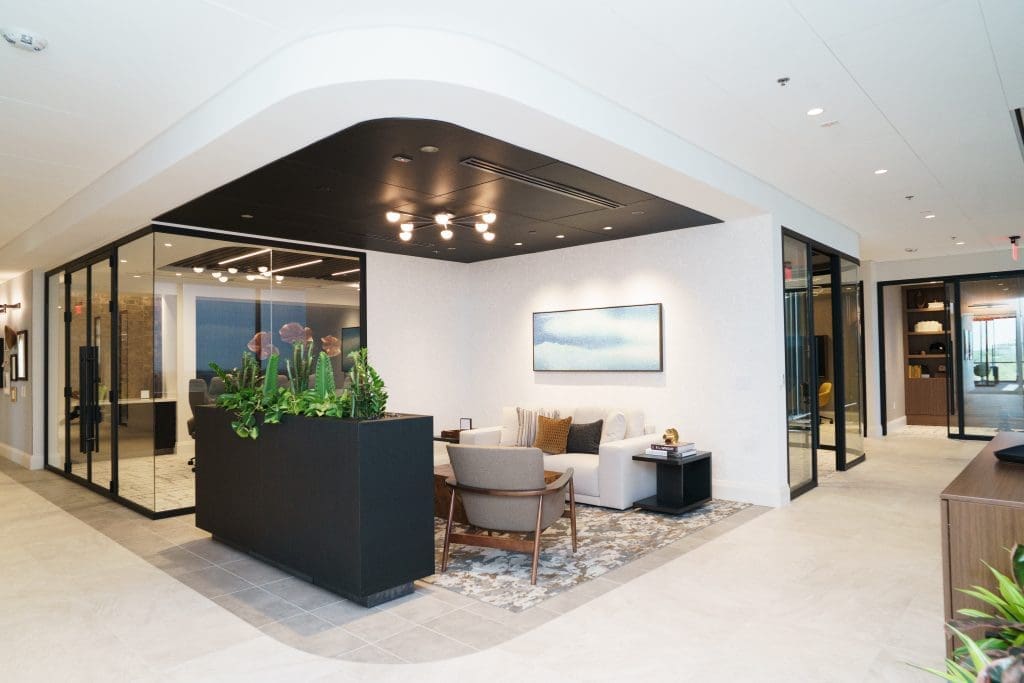 Client sitting area with two gray chairs and a white sofa, large brown planter filled with bright green plants, white walls and light grey flooring with dark brown wood panelling on the ceiling.