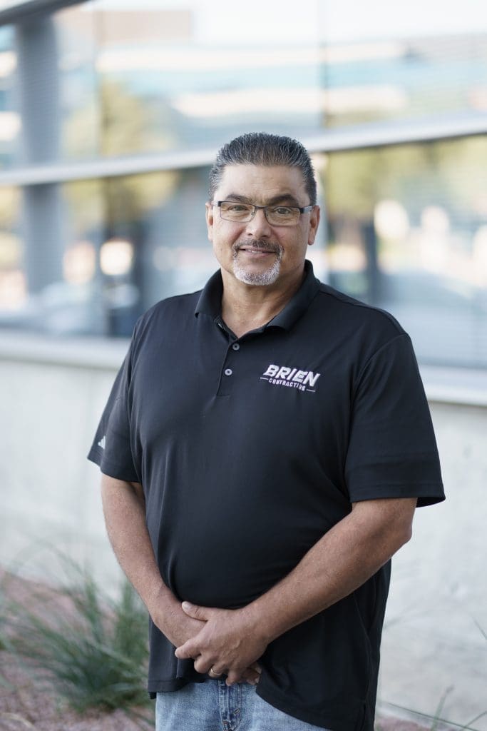 Brien Contracting Superintendent with Brien Contracting standing outside with a black polo shirt with a company logo. He has facial hair and wears glasses.