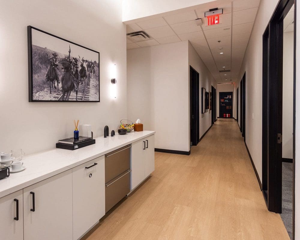 A hallway in an office with light wood floors, white walls, and a countertop with drinks and snacks—crafted by experts in general contracting services Arizona—features a black-and-white photo of people on horseback on the wall.