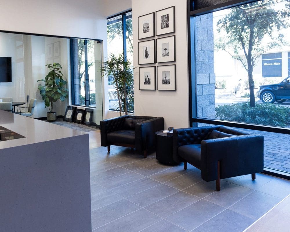 Modern office lobby with two black armchairs, a small round table, large windows, framed photos on the wall, and indoor plants. A car and trees are visible outside—ideal for businesses seeking general contracting services in Arizona.