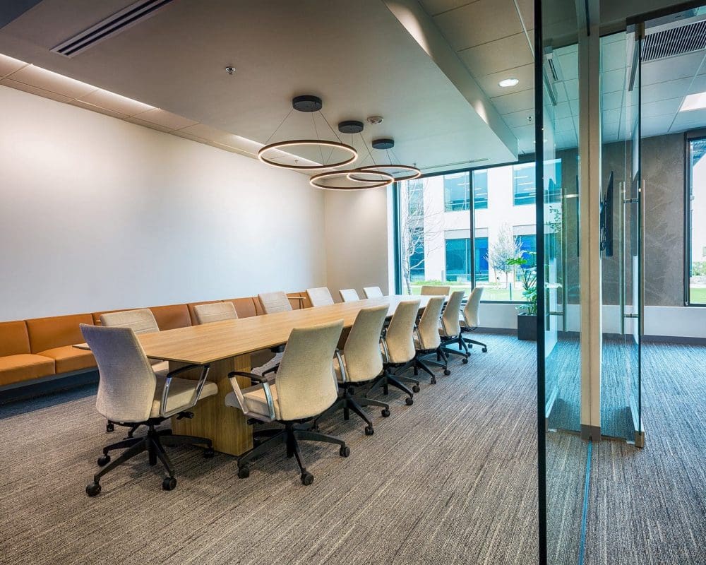 Modern conference room featuring a long wooden table, twelve office chairs, overhead circular lights, a wall-mounted couch, and glass walls—expertly crafted through general contracting services in Arizona.
