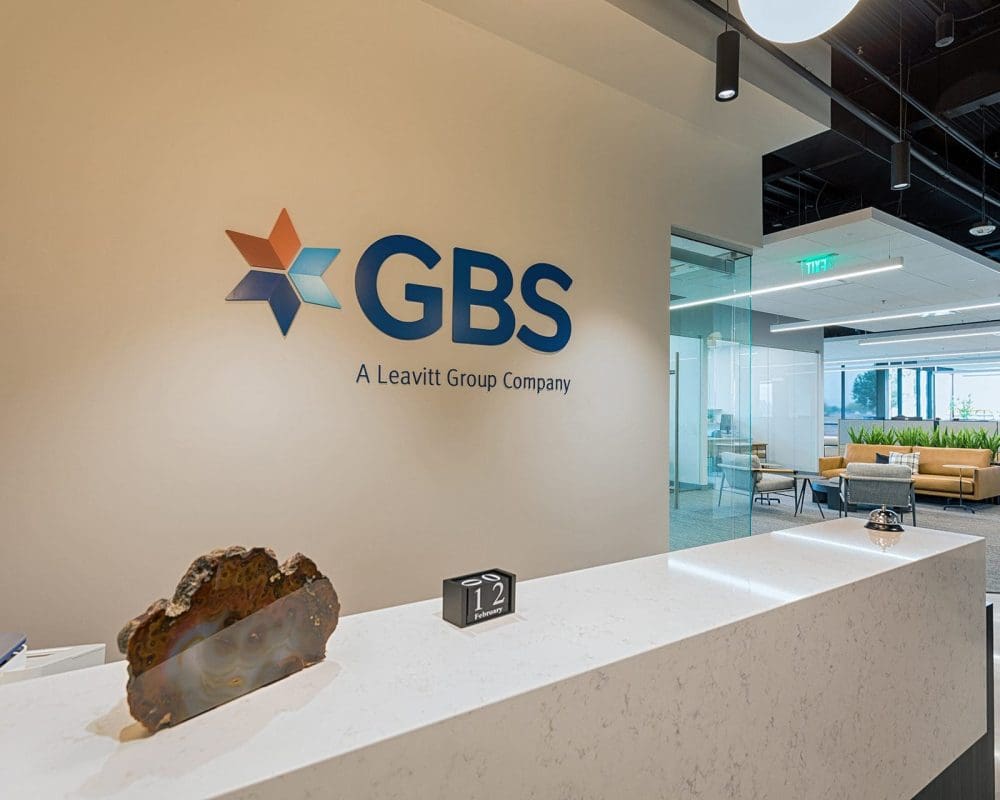 Modern office reception area with a white marble desk, decorative stone, digital clock, and GBS company logo on the wall; seating area visible in the background—showcasing expertise in general contracting services Arizona.