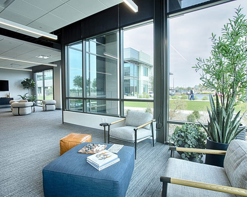 Modern office lounge area with armchairs, ottomans, coffee table with books and plants, large windows, and a view of an office building and landscaped lawn—crafted with expertise in general contracting services Arizona.