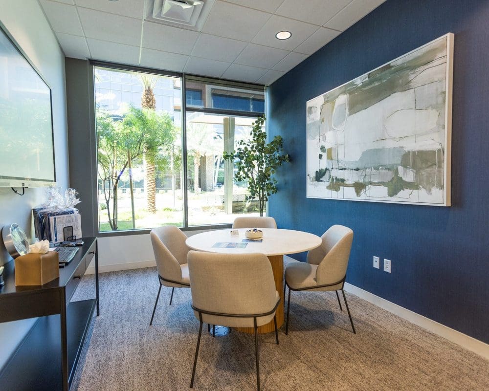 A small modern meeting room with a round table, four chairs, a large abstract painting, and a window overlooking greenery—expertly finished through professional general contracting services in Arizona.