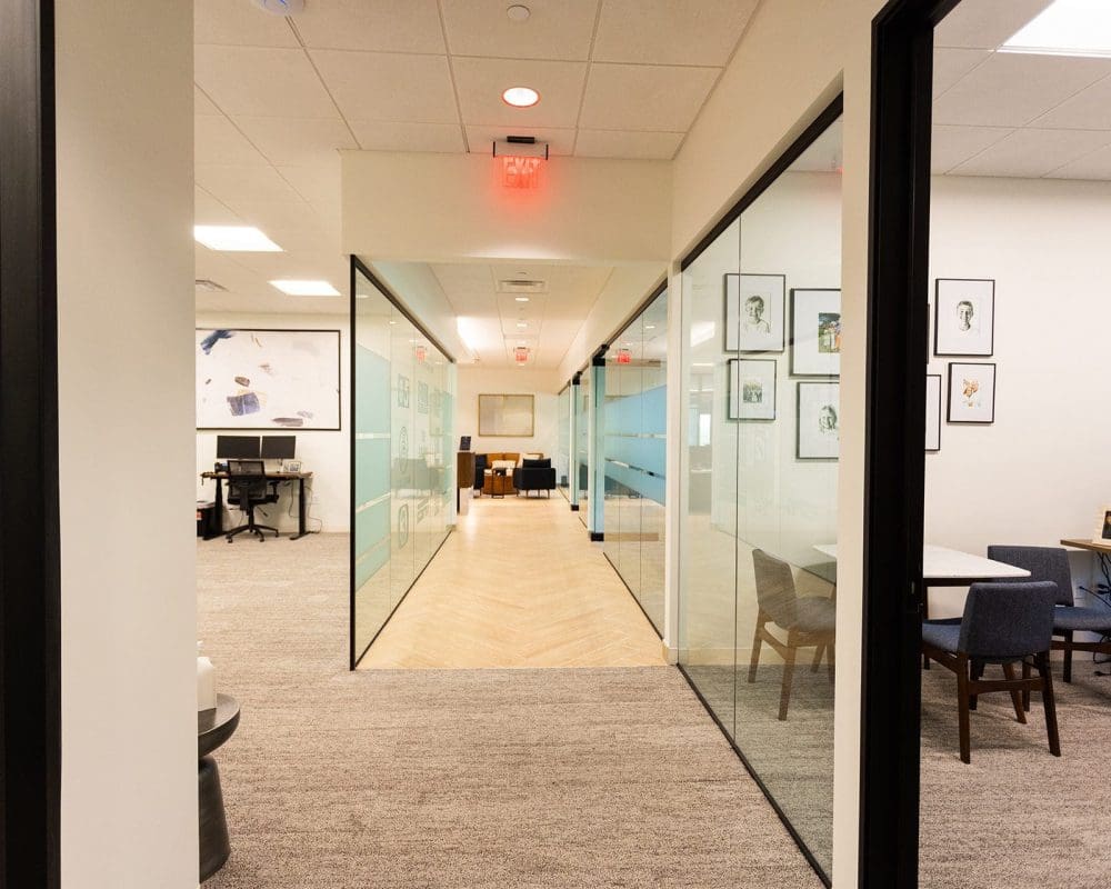 Modern office hallway featuring glass-walled conference rooms, neutral carpeting, framed art, and overhead lighting—expertly crafted through quality general contracting services in Arizona.