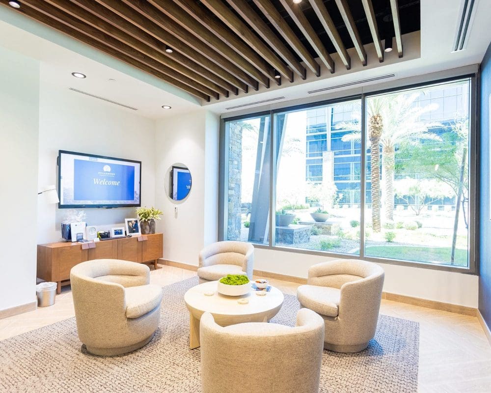 Modern office lounge with four beige chairs around a round table, large window with garden view, wall-mounted TV, and wooden ceiling accents—expertly crafted through top-tier general contracting services in Arizona.
