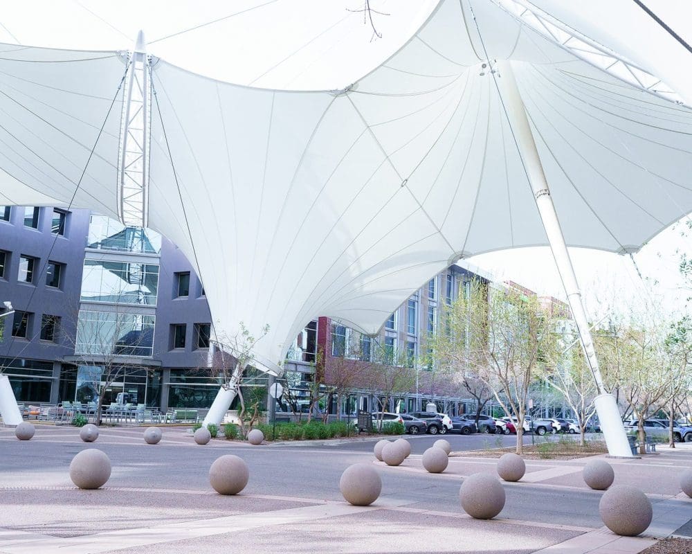 Large white shade structures cover a modern urban plaza with round concrete bollards and nearby office buildings, showcasing the quality of general contracting services Arizona businesses trust.