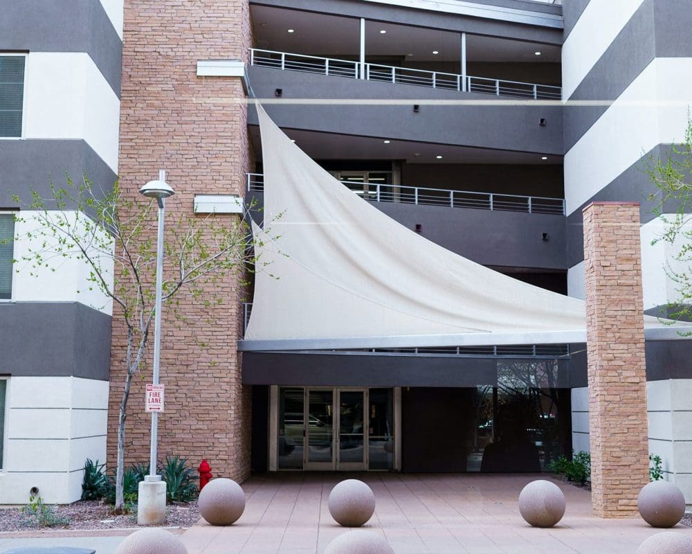 Entrance of a modern apartment building with a white sail-shaped canopy, stone accents, symmetrical windows, and round concrete bollards lining the walkway—expertly crafted by general contracting services in Arizona.