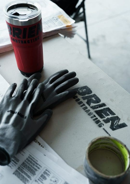 A pair of work gloves, a sealed tumbler, and a roll of tape rest on a table with the "Brien Contracting" logo—essentials for tackling commercial construction jobs in Arizona—next to printed documents.
